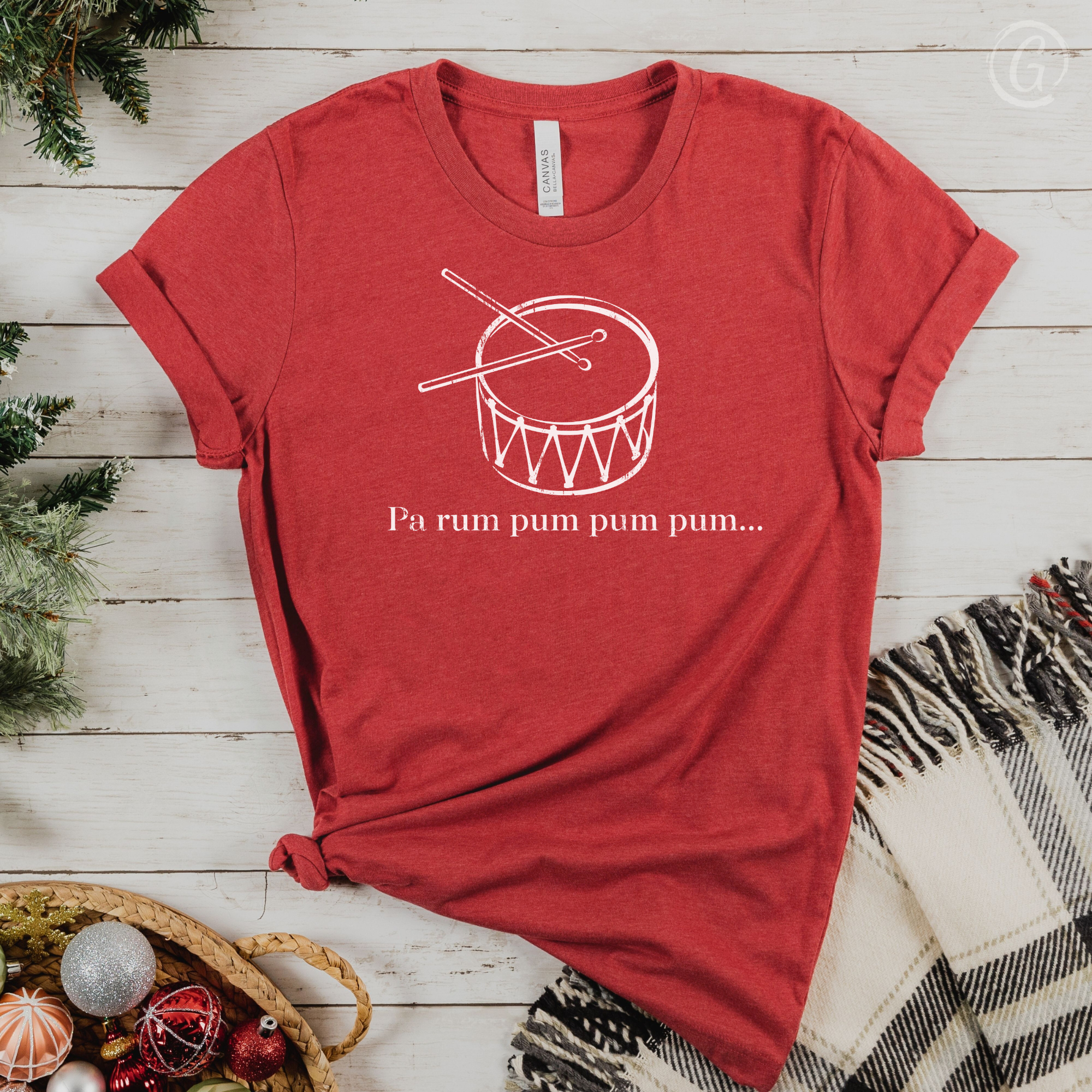 Red t-shirt with a white drum graphic and text on a wooden surface with Christmas decorations.
