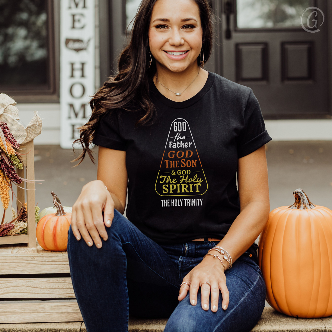 Woman wearing a black t-shirt with religious design, sitting on steps with pumpkins and autumn decorations.