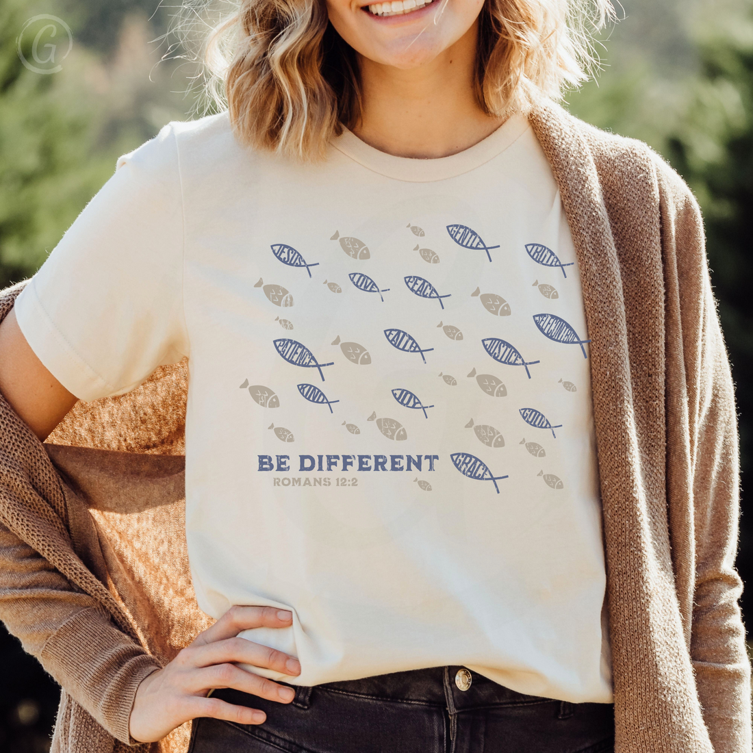 A navy-colored unisex t-shirt with a graphic design and text, displayed on a flat surface with a striped fabric, a book, and a coffee cup in the background.
