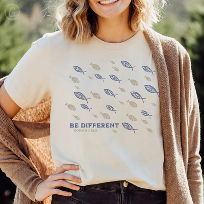 A navy-colored unisex t-shirt with a graphic design and text, displayed on a flat surface with a striped fabric, a book, and a coffee cup in the background.
